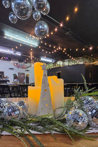 Load image into Gallery viewer, A festive holiday centerpiece on a table featuring two tall white pillar candles, green pine branches, and several mirrored disco balls, with a decorated event venue in the background.
